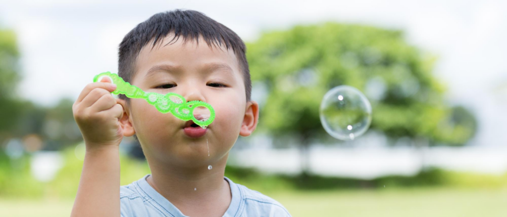 Child with a joyful expression blowing a bubble, cheeks puffed out slightly as they use their facial muscles. The iridescent bubble reflects light, adding a touch of whimsy to the moment.